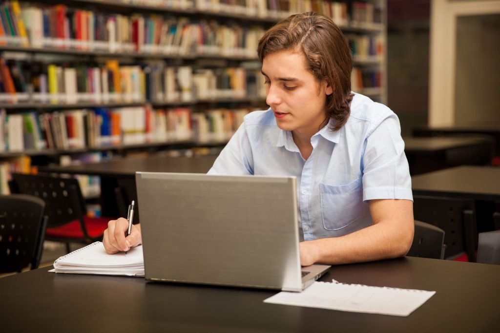 Estudante universitário fazendo anotações e usando um notebook na biblioteca