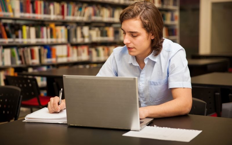 Estudante universitário fazendo anotações e usando um notebook na biblioteca
